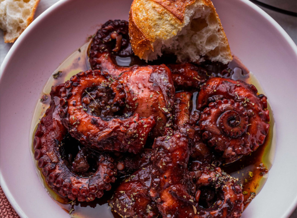 Octopus in a shallow bowl, covered in a rich red wine sauce, with a broken off piece of bread.