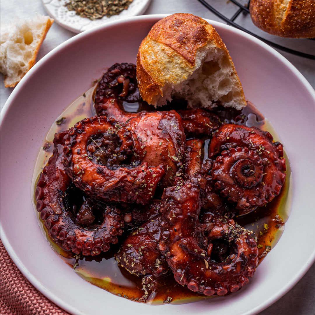 Octopus in a shallow bowl, covered in a rich red wine sauce, with a broken off piece of bread.